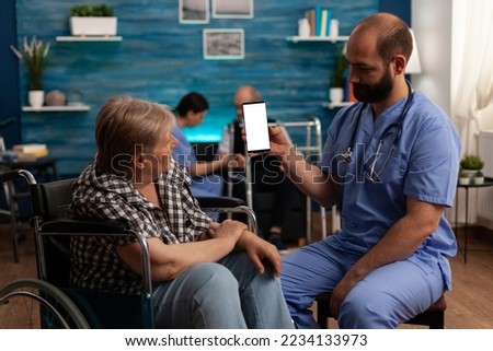 Elderly woman seated in wheelchair next to medical assistant looking at blank display on mobile device. Male nurse holding white screen on smart phone in nursing home, professional care.