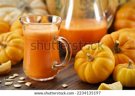 A glass cup of pumpkin juice. Pumpkins on kitchen table. Jug of pumpkin juice on background.