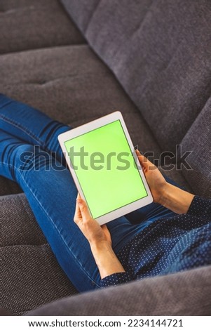 Vertical photo of unrecognizable woman lying on the couch, holding a tablet with green screen in her hands