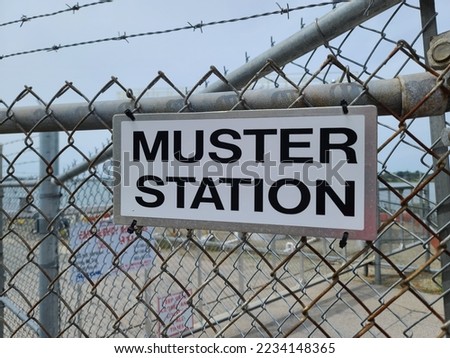 A muster station sign attached to a steel gate with barb wire above it.