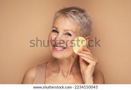 Photo of nice old woman use sponge remove make up on beige background, close up