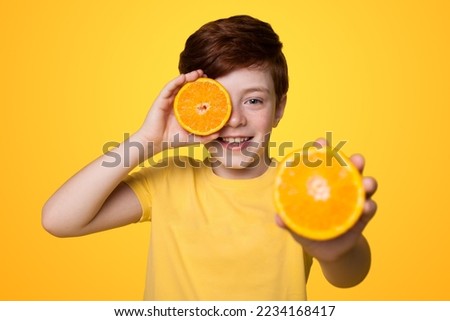 Boy holding two slices of orange in his hands, he covers his eye with one and holds the other towards the camera isolated over yellow background. Royalty-Free Stock Photo #2234168417