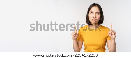 Disappointed asian woman looking, pointing fingers up with angry moody face expression, standing in yellow tshirt over white background.