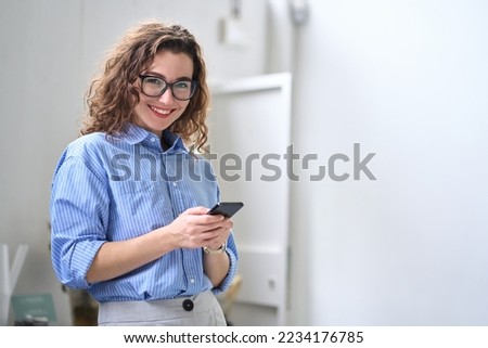 Smiling young business woman user, happy businesswoman wearing glasses holding cellular smartphone working standing in office using mobile cell phone working on cellphone looking at camera. Royalty-Free Stock Photo #2234176785