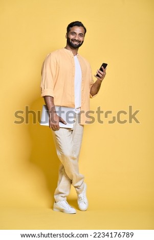 Smiling indian man user, student or employee standing isolated on yellow background holding laptop using mobile phone advertising digital products for work and learning. Full length vertical portrait.