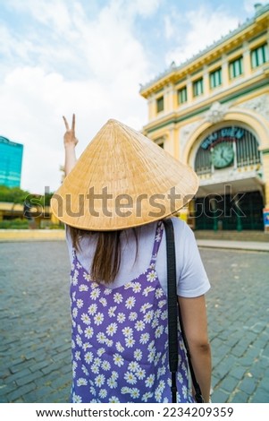 A asian traveler wearing conical hat ('Non la') taking photos in front of the Center post office in Ho Chi Minh city (Saigon), Vietnam. Travel concept.