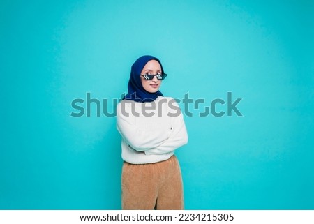 Portrait of a confident smiling Asian Muslim woman wearing a glasses, standing with arms folded and looking at the camera isolated over blue background