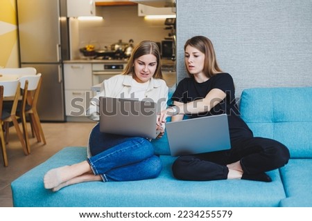 Two cute women are chatting with their friends via video call using a laptop in the living room. Friends, friendship, time together. Girlfriends are sitting on the couch and watching a movie