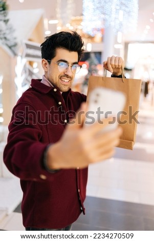 Vertical shot of positive young man taking selfie on mobile phone posing with paper bags with gifts standing in hall of celebrate shopping mall, with bright beautiful xmas decorations in Christmas eve