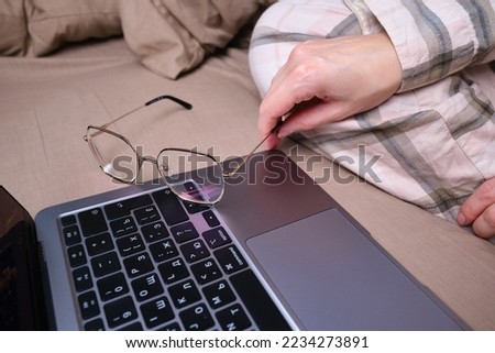 Young woman working on a laptop in glasses in bed. Glasses for vision in hands close-up. Freelance work from home