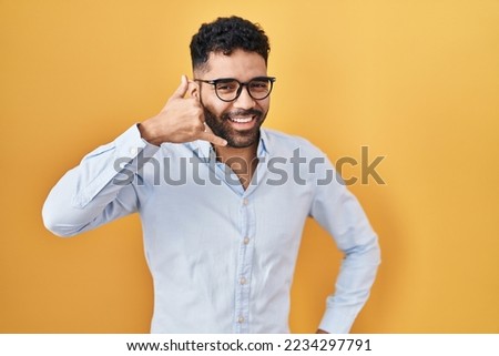 Hispanic man with beard standing over yellow background smiling doing phone gesture with hand and fingers like talking on the telephone. communicating concepts. 