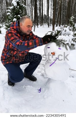 An elderly man in a red plaid jacket makes a snowman