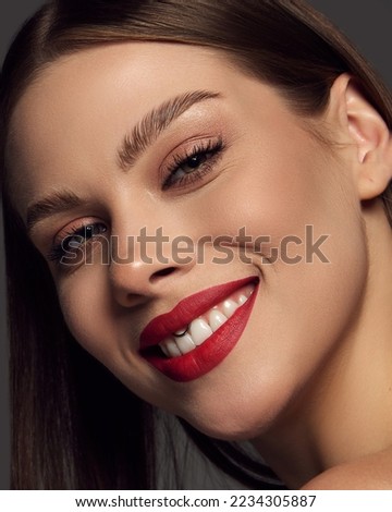 Close-up portrait of emotive beautiful young woman with red lipstick makeup smiling over dark grey background. Cosmetology and make-up products. Concept of beauty, fashion, magazine, emotions and ad.