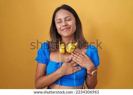 Hispanic young woman standing over yellow background smiling with hands on chest with closed eyes and grateful gesture on face. health concept. 