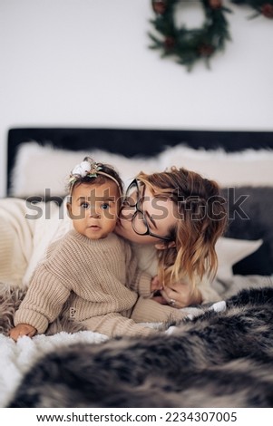 Happy caucasian woman kisses her baby daughter against background of Christmas decorations. Concept of interracial family.