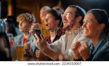 Portrait of a Happy Young Man Holding a Smartphone, Anxious About the Sports Bet on a Soccer Match. Looking at the Screen While Sitting at a Counter, Getting Very Emotional After Winning the Bet. Royalty-Free Stock Photo #2234310007