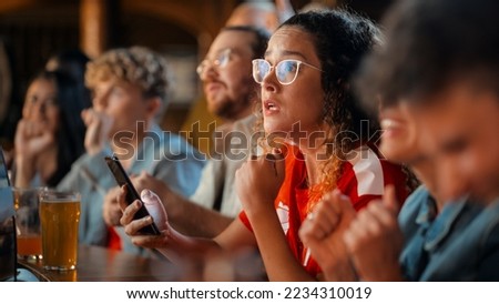 Excited Female Holding a Smartphone, Sitting at a Bar Stand. Nervous About the Sports Bet She Put on a Her Favorite Soccer Team. Joyful When Football Team Scores a Goal and She Wins the Prize. Royalty-Free Stock Photo #2234310019