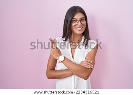 Brunette young woman standing over pink background wearing glasses with a big smile on face, pointing with hand finger to the side looking at the camera. 