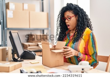 Middle age hispanic woman working at small business ecommerce looking inside box in shock face, looking skeptical and sarcastic, surprised with open mouth 