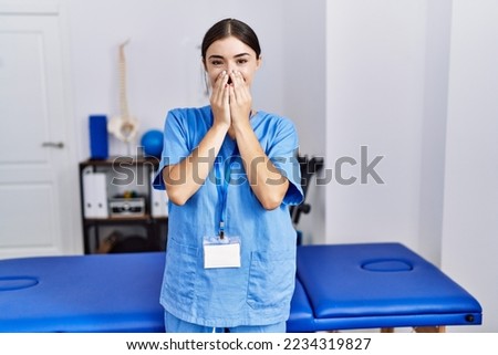 Young hispanic woman wearing physiotherapist uniform standing at clinic laughing and embarrassed giggle covering mouth with hands, gossip and scandal concept  Royalty-Free Stock Photo #2234319827