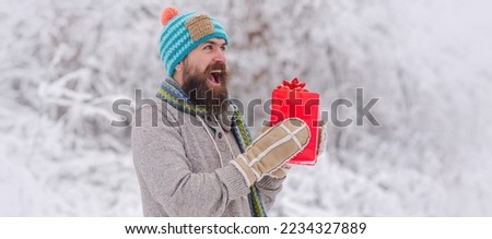 Portrait of excited man in knitted hat and winter sweater hold gift. Happy man with Christmas presents outdoor. Snowy winter background. Photo of happy Santa outdoors in snowfall carrying gifts.