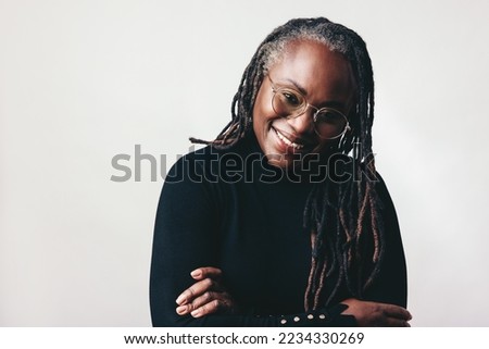 Happy professional woman smiling at the camera while wearing eyeglasses in a studio. Middle-aged woman with dreadlocks standing against a grey background. Royalty-Free Stock Photo #2234330269