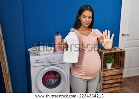 Young pregnant woman doing laundry holding detergent bottle with open hand doing stop sign with serious and confident expression, defense gesture 