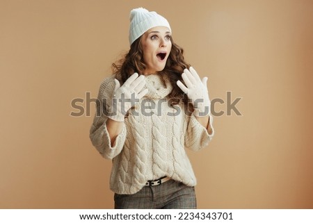 Hello winter. surprised trendy middle aged woman in beige sweater, mittens and hat on beige background.
