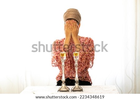 Jewish woman prays over lit Shabbat candles, covering her face with her hands. Nearby lies a religious prayer book Royalty-Free Stock Photo #2234348619