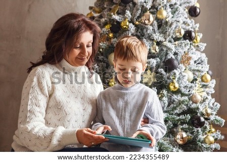 Grandmother and little boy grandson with digital tablet sitting together near beautiful decorated xmas tree at home, kid spending time with grandma during winter holidays, watching cartoons on device