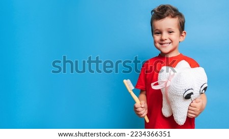 A smiling boy with healthy teeth holds a plush tooth and a toothbrush on a blue isolated background. Oral hygiene. Pediatric dentistry. Prevention of caries. A place for your text. Royalty-Free Stock Photo #2234366351