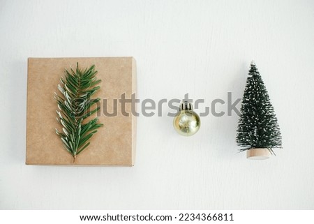 Merry Christmas! Stylish eco christmas gift box, golden bauble and tree decoration on white table flat lay. Simple craft christmas present on rustic wood. Zero waste holiday. Minimal still life