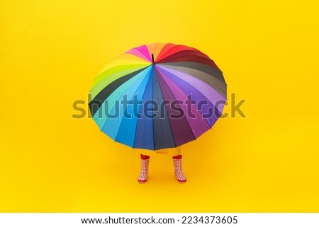 A multicolored rainbow umbrella with legs in red rubber boots on a yellow isolated background. Rainy spring, autumn weather. Legs peek out from under the umbrella.