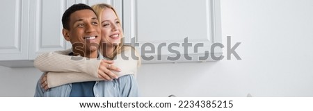 young blonde woman hugging cheerful african american boyfriend and looking away in kitchen, banner