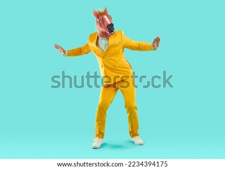 Man wearing horse head mask dancing in studio. Full length of energetic excited man wearing stylish yellow party suit and animal mask dancing spreading hands on isolated light blue studio background Royalty-Free Stock Photo #2234394175