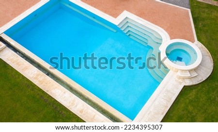 Aerial view of a blue pool with stairs to descend and climb into the water. Around marble tiles and grass. 