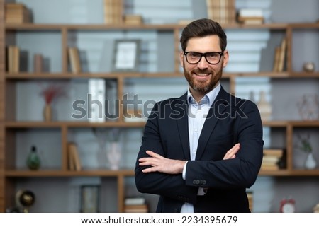 Portrait of successful mature boss, senior businessman in glasses and business suit looking at camera and smiling, man with crossed arms working inside modern office building. Royalty-Free Stock Photo #2234395679