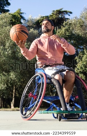 Bearded man in sports wheelchair holding basketball outside while spending time in park. Man with disability playing ball game at sports ground. Amputee sport, healthy lifestyle concept