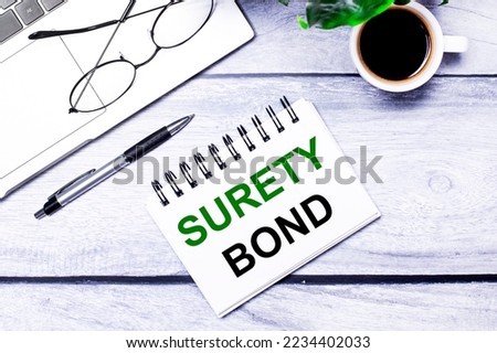 Laptop keyboard on wooden office table with coffee cup, computer mouse and notepad with SURETY BOND text. Business and finance concept. Workplace, flat lay with copy space. Royalty-Free Stock Photo #2234402033