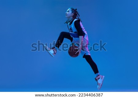 Dynamic portrait of teen girl, basketball player training, jumping with ball isolated over blue studio background in neon. Defender. Concept of professional sport, active lifestyle, hobby, team game Royalty-Free Stock Photo #2234406387