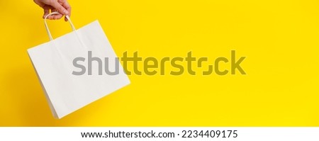 Female hand holding white blank shopping bag on yellow background. White craft blank paper bag in hand. Discount, shopping and ecology concept. Black Friday sale banner Royalty-Free Stock Photo #2234409175