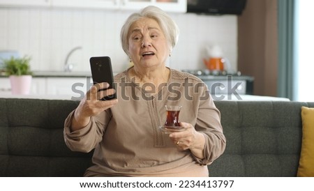 Happy old grandmother holding mobile phone is very surprised to hear that she is making video call with loved ones while drinking tea. An elderly woman learning to use modern technology device.