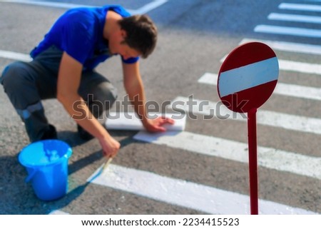 Road service worker sits on his lap and paints striped pedestrian crossing on asphalt with brush. Markings for pedestrians on roadway. Safe transition for people. Real workflow..