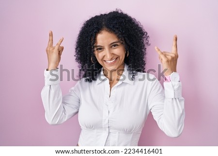 Hispanic woman with curly hair standing over pink background shouting with crazy expression doing rock symbol with hands up. music star. heavy music concept. 