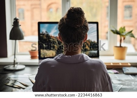 Young woman working on computer in home office and editing photos, view from back