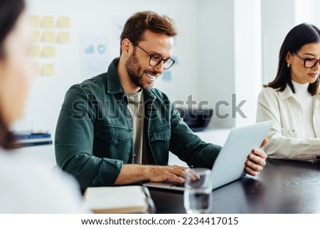 Designer using a laptop during a meeting in an office. Happy business man reading an email while sitting in a boardroom with his colleagues. Royalty-Free Stock Photo #2234417015