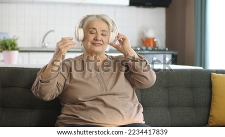 Colorful and funny moments of an old woman listening to music with headphones on her sofa. Funny old woman with headphones singing and dancing. Mature cheerful woman enjoying life after retirement.