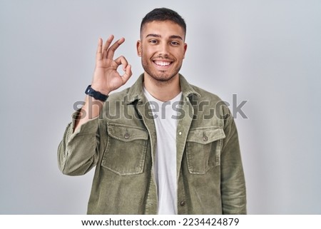 Young hispanic man standing over isolated background smiling positive doing ok sign with hand and fingers. successful expression. 