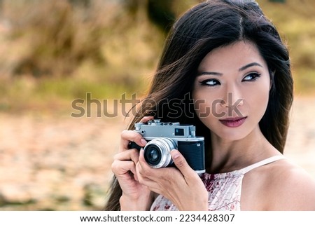 Vintage style portrait of beautiful Asian Chinese girl or young woman wearing summer dress outside in green environment taking pictures or photographs in Autumn or Fall with a retro camera
