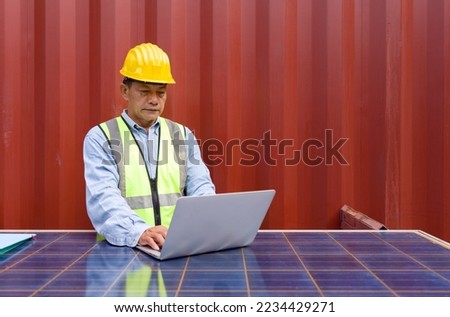 Senior contractor in safety vest and yellow hardhat typing on laptop computer keyboard. Check out the delivery schedule planning for the next shipment. He use solar cell panel as a table.